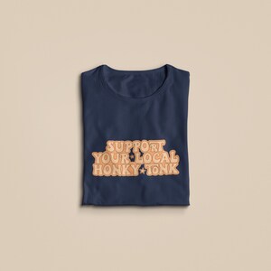 Puede incluir: Una camiseta azul marino con el texto "Support Your Local Honky Tonk" en naranja.