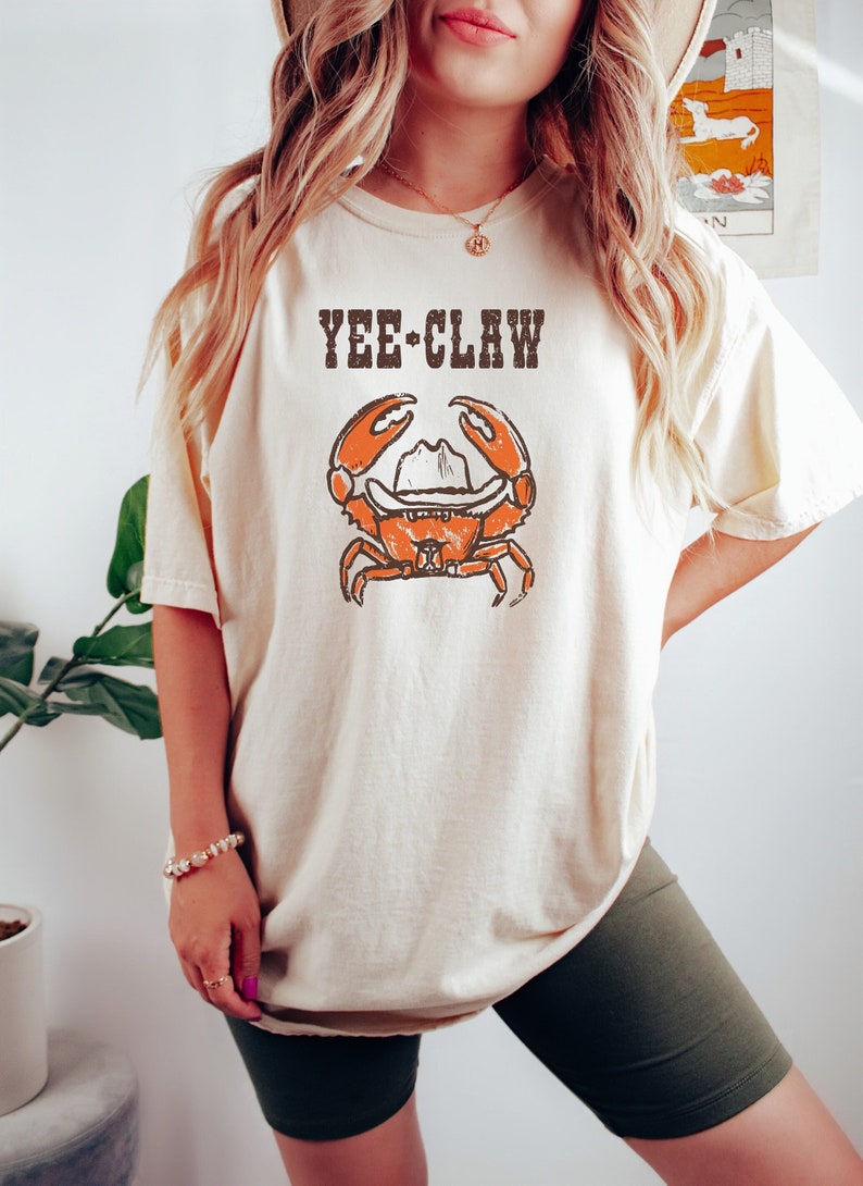 May include: Cream-colored t-shirt featuring a distressed graphic of an orange crab wearing a cowboy hat. The text "YEE-CLAW" is printed above the crab. The shirt is a relaxed fit.