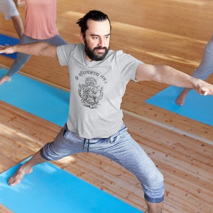 May include: A man in a grey t-shirt with a black and white graphic of a deity and the text "ॐ गणानायक नमः" is doing a yoga pose on a blue mat. He is in a yoga studio with wooden floors.