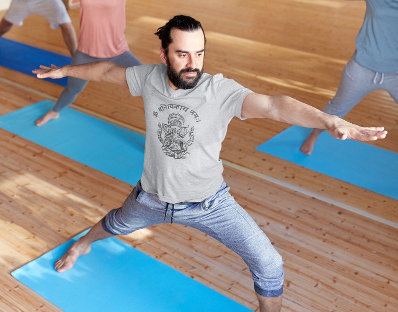 May include: A man in a grey t-shirt with a black and white graphic of a deity and the text "ॐ गणानायक नमः" is doing a yoga pose on a blue mat. He is in a yoga studio with wooden floors.