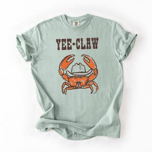 May include: A light green t-shirt featuring the text "YEE-CLAW" above a cartoon crab wearing a cowboy hat. The crab is orange with brown details. The shirt has rolled-up sleeves.