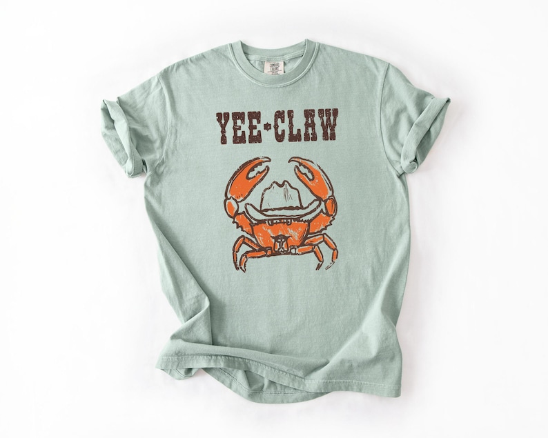 May include: A light green t-shirt featuring the text "YEE-CLAW" above a cartoon crab wearing a cowboy hat. The crab is orange with brown details. The shirt has rolled-up sleeves.