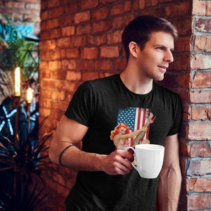 May include: A man wearing a black t-shirt with a vintage-style image of a woman in a red, white, and blue outfit holding a red, white, and blue flag. The man is holding a white coffee mug.
