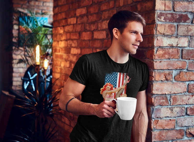 May include: A man wearing a black t-shirt with a vintage-style image of a woman in a red, white, and blue outfit holding a red, white, and blue flag. The man is holding a white coffee mug.