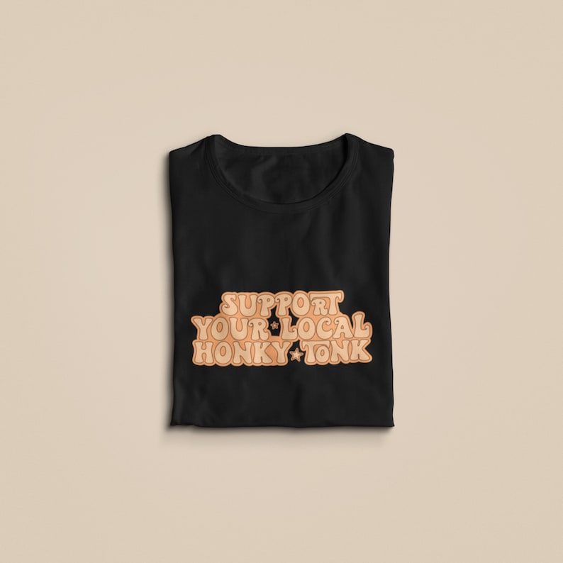 Puede incluir: Camiseta negra con el texto "Support Your Local Honky Tonk" en una fuente naranja retro.