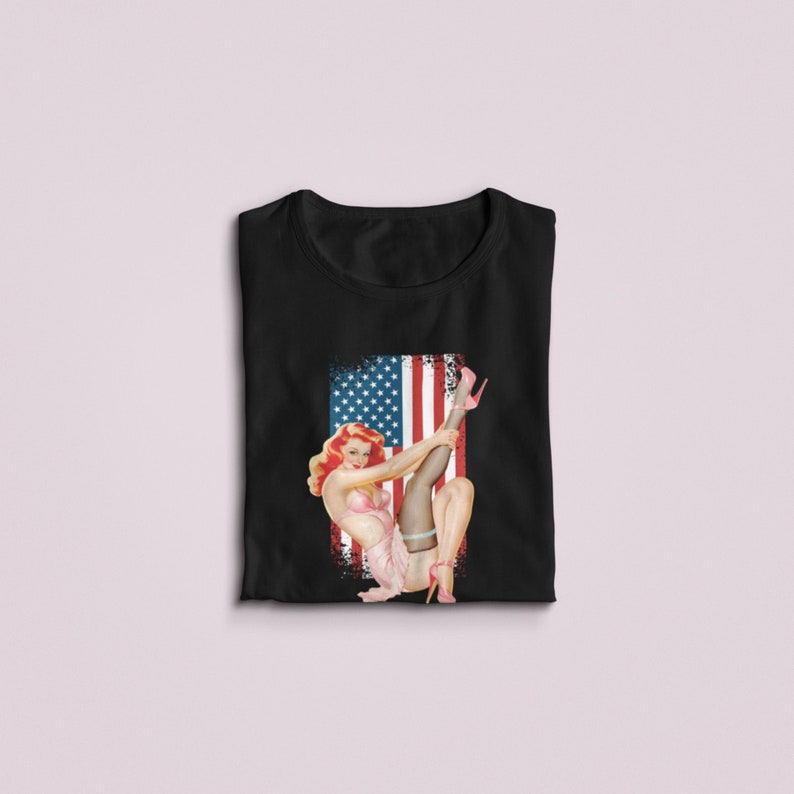 May include: Black t-shirt with a vintage illustration of a woman in a pink dress and stockings, posing in front of an American flag.