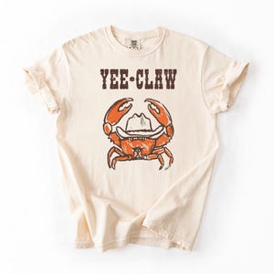 May include: Cream-colored t-shirt with the text "YEE-CLAW" above a cartoon crab wearing a cowboy hat. The crab is orange with brown details. The shirt has short sleeves and a crew neck.