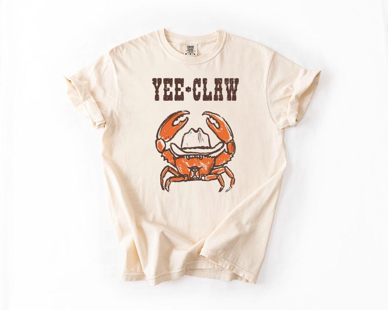 May include: Cream-colored t-shirt with the text "YEE-CLAW" above a cartoon crab wearing a cowboy hat. The crab is orange with brown details. The shirt has short sleeves and a crew neck.