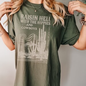 Puede incluir: Camiseta verde con un estampado gr&aacute;fico blanco que dice "Raisin Hell with the Hippies and Cowboys" y presenta un cactus con el texto "Can Not Be Tamed" y "Yee Haw".