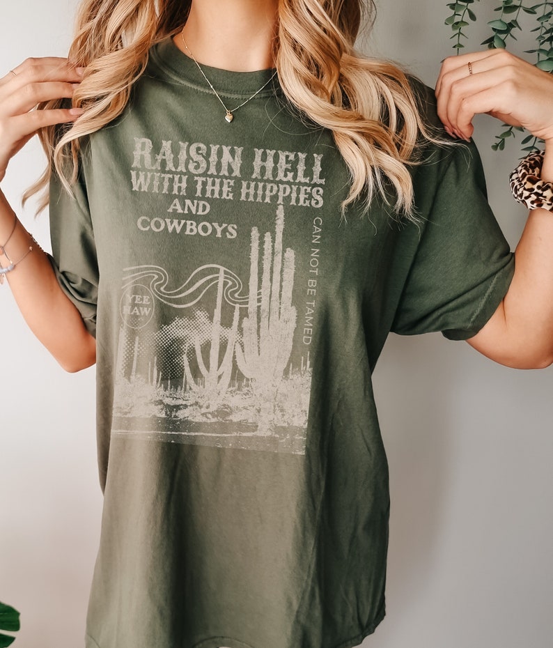 Puede incluir: Camiseta verde con un estampado gr&aacute;fico blanco que dice "Raisin Hell with the Hippies and Cowboys" y presenta un cactus con el texto "Can Not Be Tamed" y "Yee Haw".