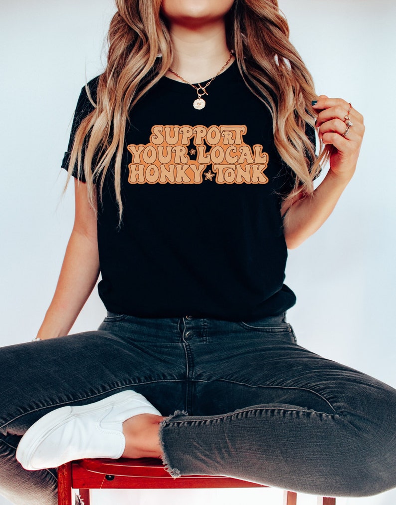 Puede incluir: Una camiseta negra con el texto "Support Your Local Honky Tonk" en una fuente naranja retro.