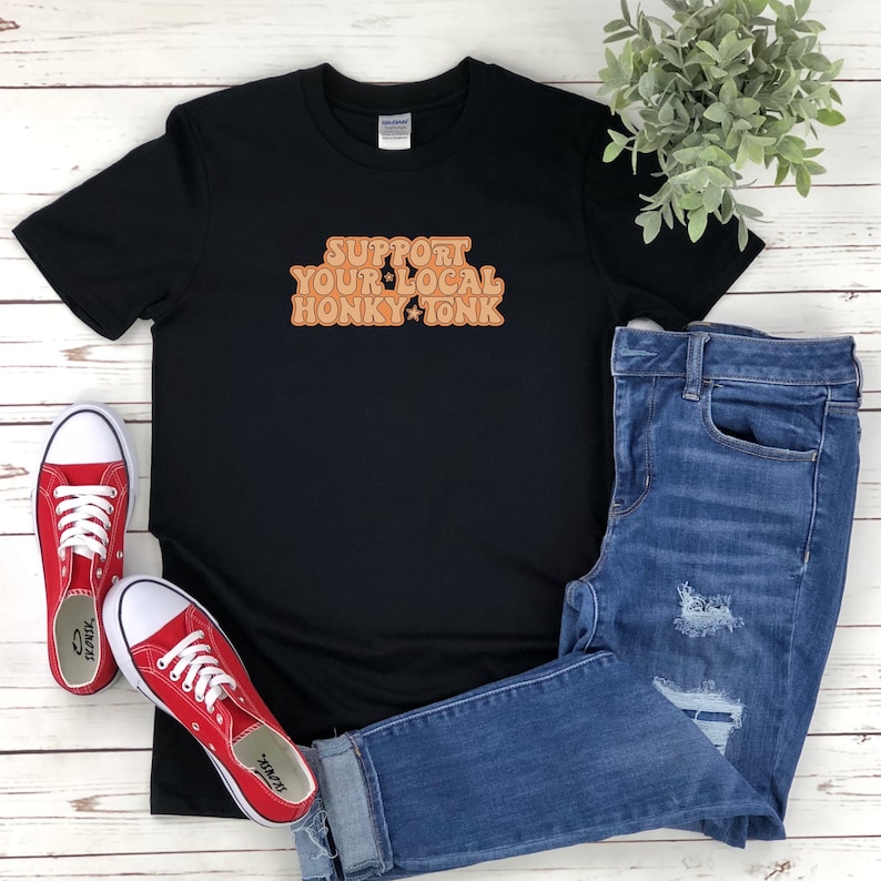Puede incluir: Camiseta negra con el texto "Support Your Local Honky Tonk" en naranja con una estrella en el medio de la palabra "Honky".