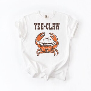 May include: White t-shirt with a distressed graphic of an orange crab wearing a cowboy hat. The text "YEE-CLAW" is printed above the crab. The shirt is made of a soft material.