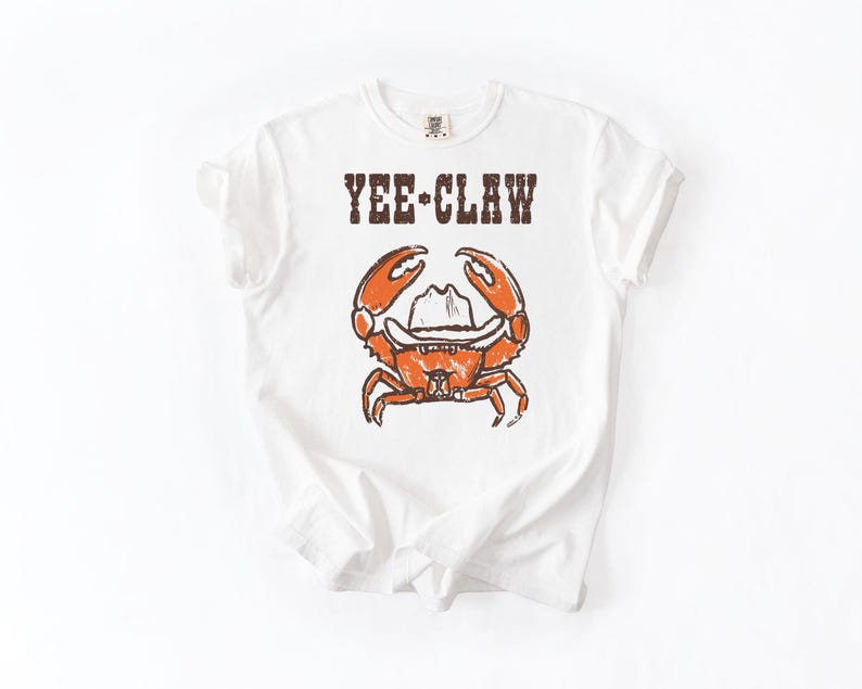 May include: White t-shirt with a distressed graphic of an orange crab wearing a cowboy hat. The text "YEE-CLAW" is printed above the crab. The shirt is made of a soft material.