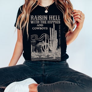 Puede incluir: Camiseta negra con un estampado gr&aacute;fico de cactus y el texto "Raisin Hell with the Hippies and Cowboys Can Not Be Tamed" y "Yee Haw" en una fuente de estilo vintage.