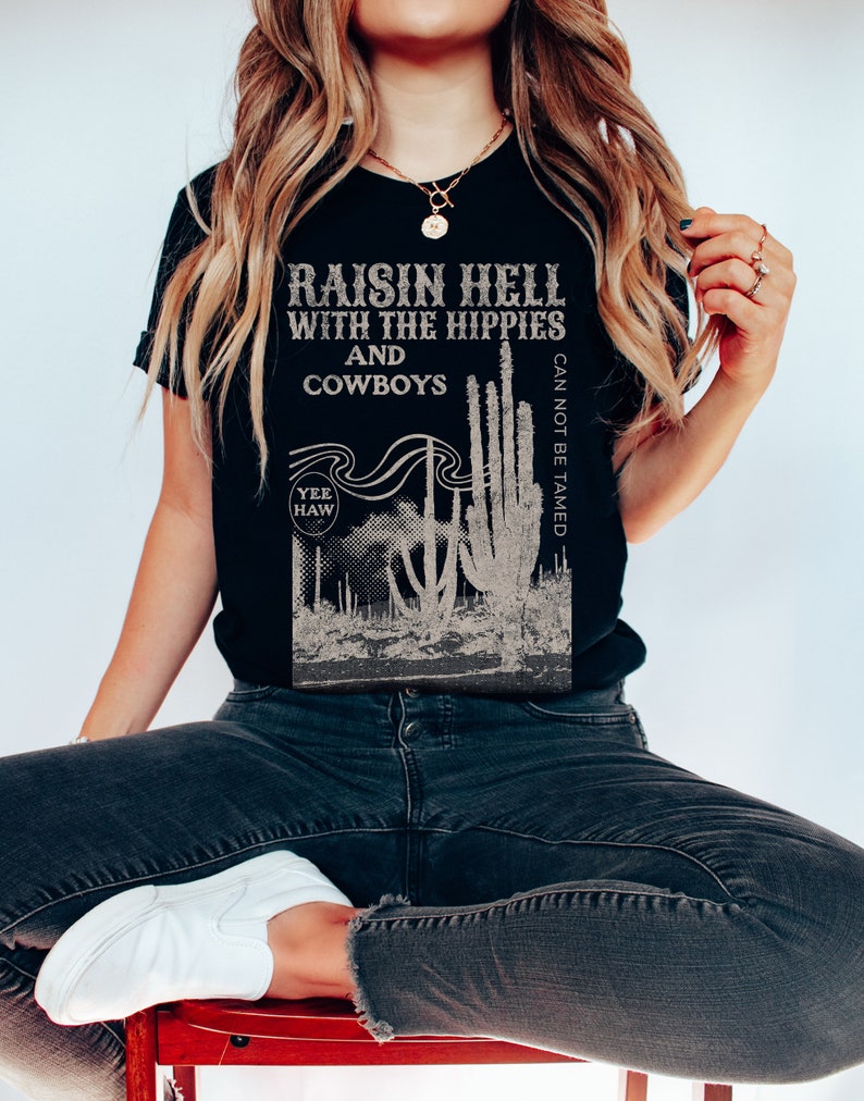 Puede incluir: Camiseta negra con un estampado gr&aacute;fico de cactus y el texto "Raisin Hell with the Hippies and Cowboys Can Not Be Tamed" y "Yee Haw" en una fuente de estilo vintage.