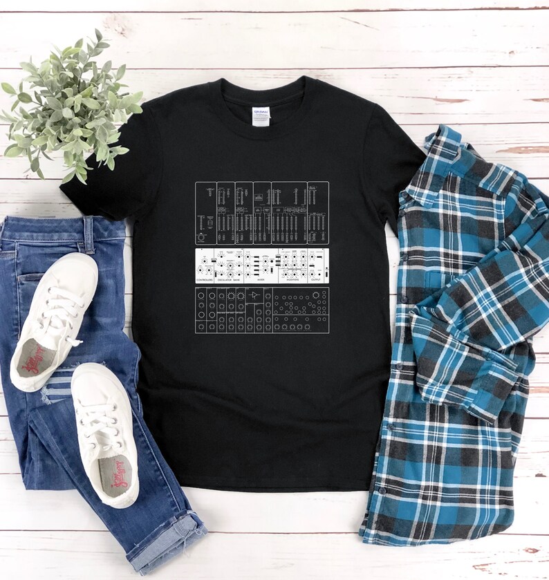 May include: A black t-shirt with a white graphic of a synthesizer panel. The panel has knobs, buttons, and sliders. The t-shirt is laid out on a white wooden surface with a pair of blue jeans and a blue plaid shirt.