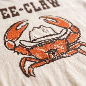 May include: Cream-colored t-shirt featuring a distressed orange crab wearing a cowboy hat. The crab design is outlined in dark brown, with the words "EE-CLAW" printed above in the same brown color. The shirt has a vintage, worn look.