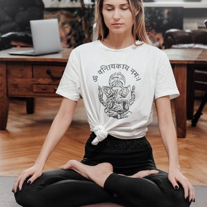 May include: A woman sits in a yoga pose on a purple yoga mat with her eyes closed. She is wearing a white t-shirt with a black and white image of Ganesha and the text "ॐ वनिनायकाय नमः". She is also wearing black leggings.