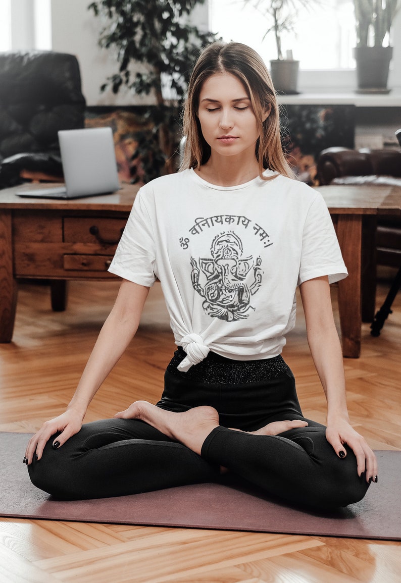 May include: A woman sits in a yoga pose on a purple yoga mat with her eyes closed. She is wearing a white t-shirt with a black and white image of Ganesha and the text "ॐ वनिनायकाय नमः". She is also wearing black leggings.
