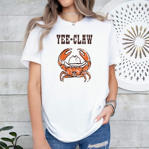 May include: White t-shirt featuring a distressed graphic of an orange crab wearing a cowboy hat, with the text "YEE CLAW" above. The design has a vintage, worn look. The shirt is a casual, everyday wear item.
