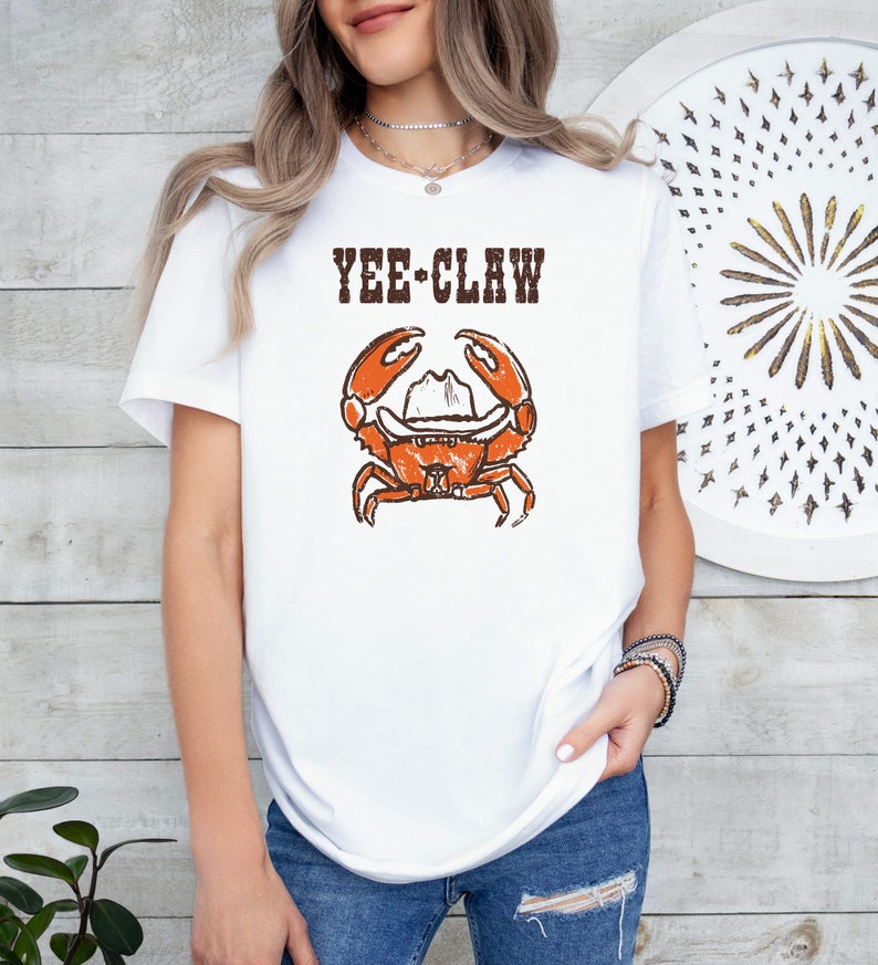May include: White t-shirt featuring a distressed graphic of an orange crab wearing a cowboy hat, with the text "YEE CLAW" above. The design has a vintage, worn look. The shirt is a casual, everyday wear item.