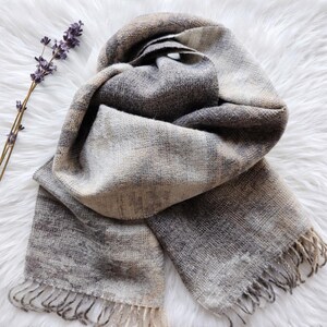 May include: A grey, beige, and brown woven scarf with fringed edges. The scarf is draped on a white, fluffy surface, with a sprig of lavender in the upper left corner. The scarf has a soft, textured appearance.