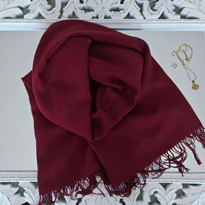 May include: A deep red scarf with fringed edges lies on a mirror. A gold necklace with a pendant and stud earrings are also on the mirror. The mirror is set in a white, ornate, carved frame.