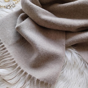 Może przedstawiać: Beżowy szalik z frędzlami, narzucony na białą, puszystą powierzchnię. Szalik wykonany jest z miękkiego, tkanego materiału. Naszyjnik i kolczyki są widoczne w tle.