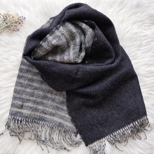 May include: A dark grey scarf with a textured weave and fringe detail. The scarf has a reversible design, with a patterned side and a solid side. The fringe is silver and adds a touch of elegance to the accessory.