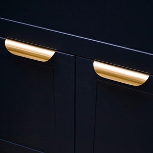 May include: Two gold-toned cabinet pulls are mounted on dark blue cabinet doors. The pulls are curved and have a smooth, polished finish.