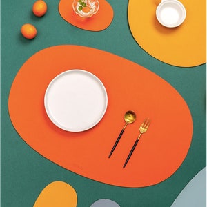 May include: A set of six colorful placemats in orange, yellow, and gray. The placemats are oval shaped and have a smooth, textured surface. The placemats are perfect for adding a touch of color and style to any dining table.