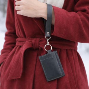 Peut inclure: Un porte-cartes en cuir noir avec une dragonne. Le porte-cartes est rectangulaire avec une bordure cousue et une poche sur le devant. La dragonne est en cuir noir avec un fermoir argenté.