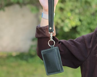 May include: A black leather cardholder with a matching wrist strap. The cardholder has a pocket and is stitched with light green thread. The strap is attached with a silver-colored clasp and a metal ring. The background is a blurred green.
