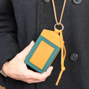 May include: A two-tone ID holder in orange and teal, with a clear window. The holder is attached to a tan lanyard with a metal ring. The holder is being held by a person wearing a dark coat.