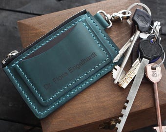 May include: Teal leather key holder with a zipper and a card slot, featuring the engraved name "Dr. Floris Engelhardt." The key holder is attached to a silver keychain with several keys. The stitching is a lighter teal color.