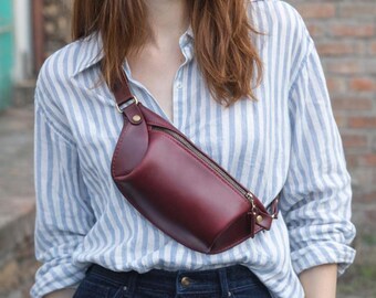 May include: A burgundy leather bum bag with a gold zip and hardware. The bag is worn across the body. The person is wearing a blue and white striped button-down shirt and dark blue jeans.