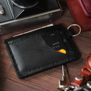 Puede incluir: Tarjetero de cuero negro con un llavero, que contiene una tarjeta de crédito y algo de efectivo. El tarjetero tiene un borde cosido y un diseño minimalista. Una cámara vintage está en el fondo.