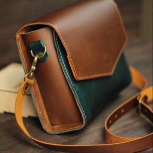 May include: A brown and teal leather crossbody bag with a flap closure. The bag features a tan adjustable strap and gold-colored hardware. The bag is made of stitched leather.