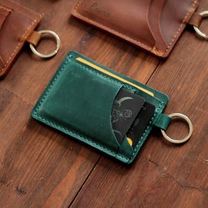 Könnte beinhalten: Ein grüner Lederkartenhalter mit Schlüsselring, der zwei schwarze Karten enthält. Der Kartenhalter ist rechteckig mit einer Tasche für Karten. Weitere braune Lederkartenhalter sind im Hintergrund zu sehen.
