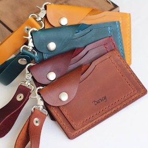 May include: A collection of handmade leather cardholders in various colors, including brown, teal, and burgundy. Each wallet features a snap closure and a wrist strap with a metal clasp. The word "Darcy" is embossed on one wallet.