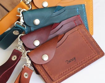 May include: A collection of handmade leather cardholders in various colors, including brown, teal, and burgundy. Each wallet features a snap closure and a wrist strap with a metal clasp. The word "Darcy" is embossed on one wallet.