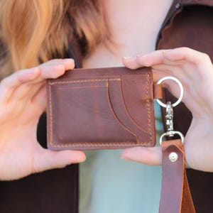 Può includere: Un porta carte in pelle marrone con più fessure per carte e un portachiavi. Il porta carte è rettangolare con cuciture visibili sui bordi. Un cinturino in pelle marrone è attaccato al portachiavi.