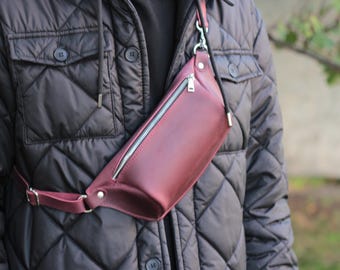 May include: A burgundy leather fanny pack with a silver zip and adjustable strap. The bag is worn across the body, highlighting its sleek design and practical functionality. The bag is made of high-quality leather.