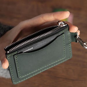 May include: A dark green leather card holder featuring a top zip closure with a silver-toned metal pull. The exterior includes a front pocket containing a black card with the text monobank and Universal Bank. A silver metal lobster clasp is attached.