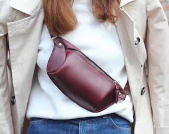 May include: A burgundy leather bum bag with a silver zip, worn across the chest. The bag is a deep red colour and appears to be made of smooth leather. The background includes a white sweater, blue jeans, and a beige trench coat.