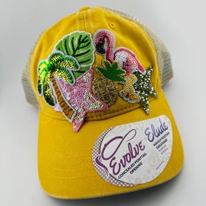 May include: Mustard yellow baseball cap with a mesh back. The cap is decorated with sequined tropical designs, including a flamingo, palm tree, and star. The tag reads "Evolve Elude" with "Concealed Ponytail Opening".