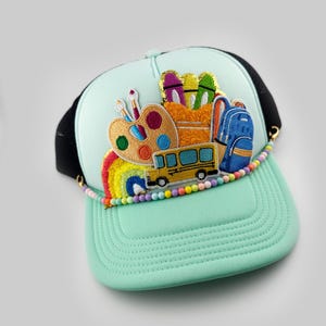 May include: A light blue trucker hat with a black mesh back. The hat features colorful embroidered patches of school supplies, including a paint palette, crayons, a backpack, and a school bus. The hat also has a rainbow and colorful beads around the brim.
