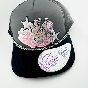 May include: Gray and black trucker hat with a silver chain detail. The hat has several embroidered patches, including a pink boot, a peace sign, and a star. The hat also has a label that says "Evolve Concealed Ponytail Opening" and "Elude Makeup Resistant Sweatband Breathable & Wicking".