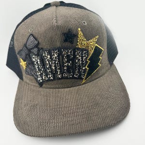 May include: A brown corduroy and black mesh baseball cap with a black and gold embroidered design featuring the word "NEW" and stars.